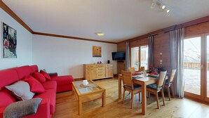 A living room with a red sofa, wooden coffee table, and a dining area with a wooden table and chairs. A TV is mounted on the wall, and there is a window with curtains.