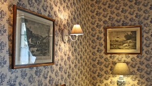 A room with patterned wallpaper, a framed picture, a lamp, a chair, and a table with a vase. - Logis Les Remparts (Bayeux)