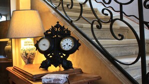 A wooden table with a clock and a lamp, next to a staircase with ornate black railings. - Logis Les Remparts (Bayeux)