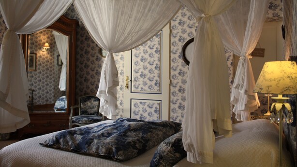A bedroom with a canopy bed, a dresser, a chair, and a lamp. - Logis Les Remparts (Bayeux)