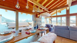 Un interior de madera con una mesa de comedor lista para una comida, un sofá y ventanas amplias que ofrecen vista a la montaña.