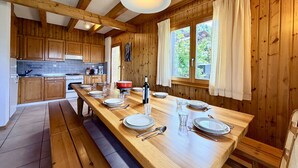 Une cuisine en bois avec une longue table à manger dressée pour quatre personnes, un poêle et une fenêtre avec des rideaux.