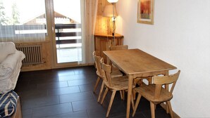 Une table à manger en bois, avec des chaises, une lampe et un radiateur, dans une pièce d’où l’on peut voir un bâtiment à travers une fenêtre.