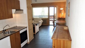 Une cuisine compacte équipée d’un évier, d’une cuisinière et de meubles de rangement en bois. On y trouve également un coin repas avec une table et des chaises, et une vue sur l’extérieur grâce à une porte coulissante.