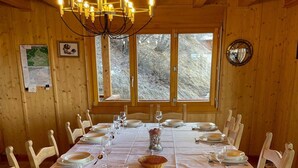 En spisestue med et langt bord dækket til et måltid, træstole, en lysekrone og vinduer med udsigt til træer.