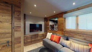 Een moderne woonkamer met een grijze bank, houten paneelwanden, een wandgemonteerde televisie en een groot raam met zonwering.