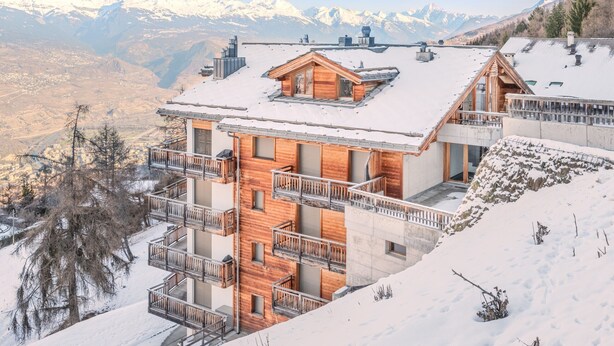 A multi-story wooden chalet with balconies, set against a snowy mountain backdrop.