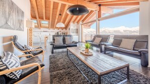 A spacious living area with a high wooden ceiling, a large window offering a mountain view, a cozy seating arrangement, and a kitchen in the background.