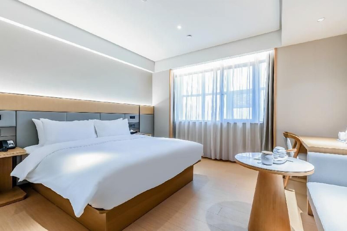 A hotel room with a large bed, a bedside table, a desk with a chair, and a window with curtains.