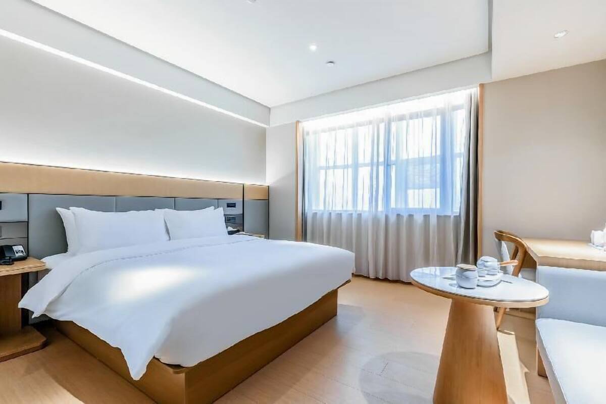 A hotel room with a large bed, a bedside table, a desk with a chair, and a window with curtains.