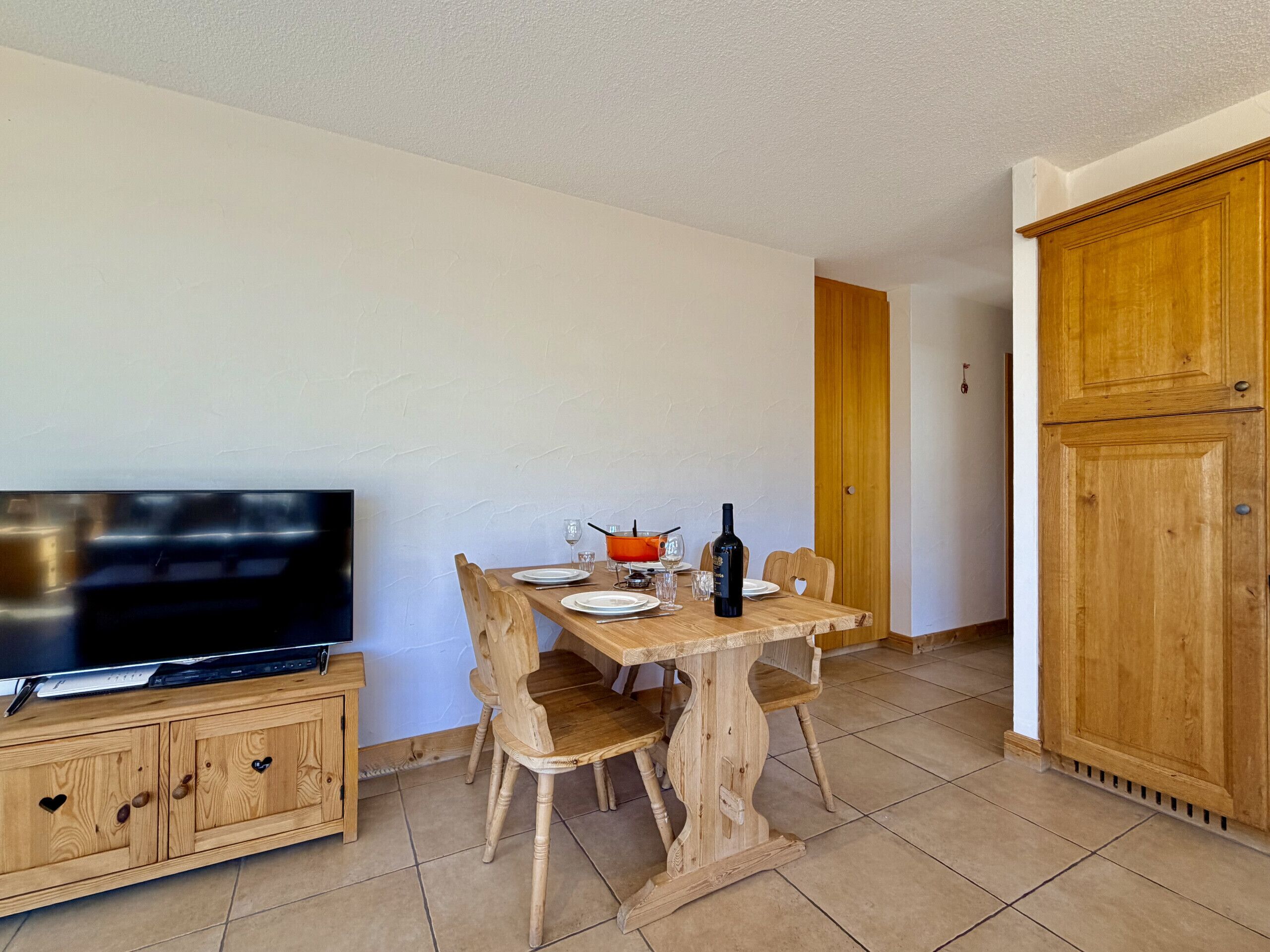 Een eethoek met een houten tafel, stoelen, een televisie op een kast en een fles wijn.