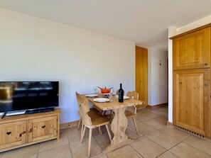 Un comedor con mesa de madera, sillas, televisor en un mueble y una botella de vino.