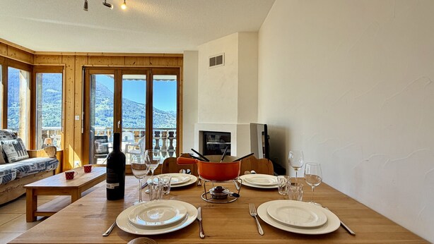 Comedor con una mesa de madera dispuesta para cuatro, un sofá y vistas a montañas a través de amplias ventanas.