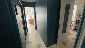 Un couloir étroit avec une porte sur la droite, qui donne sur une pièce contenant un lit et une chaise.