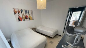 Une petite chambre propre, avec deux lits, une œuvre d’art accrochée au mur, un ventilateur sur pied et une fenêtre.