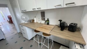 Une cuisine moderne avec des meubles blancs, un plan de travail en bois, deux tabourets de bar, un ventilateur, une radio, une machine à café, un grille-pain et un évier.