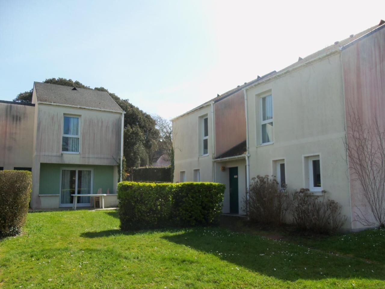 A residential building with a flat roof, a small front yard with a hedge, and a visible door.