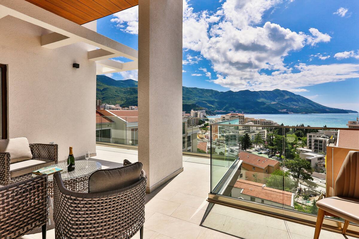 A balcony with wicker furniture, a bottle of champagne, and a glass. It overlooks a coastal city with mountains in the distance.