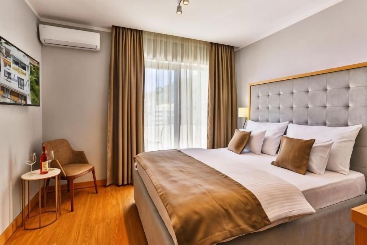 A hotel room with a large bed, a bedside table, a chair, a small table with a bottle, and a window with curtains.