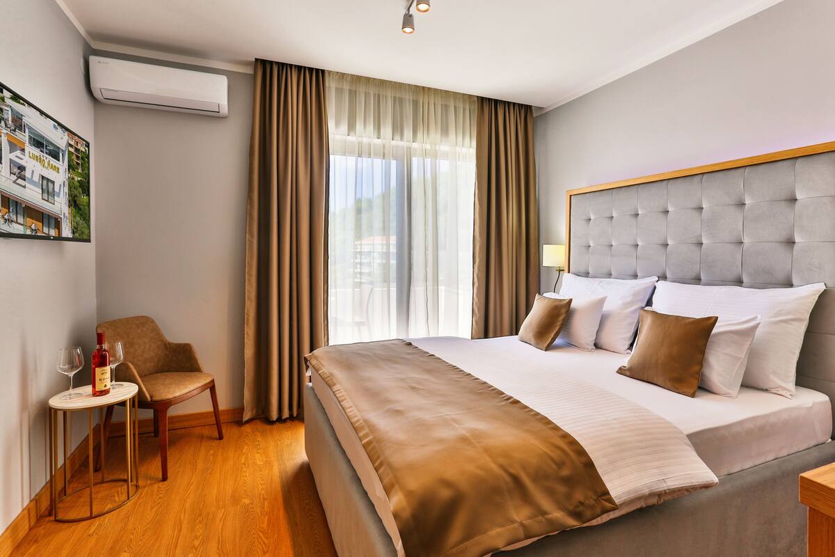 A hotel room with a large bed, a chair, a small table with a bottle and glasses, and a window with curtains.