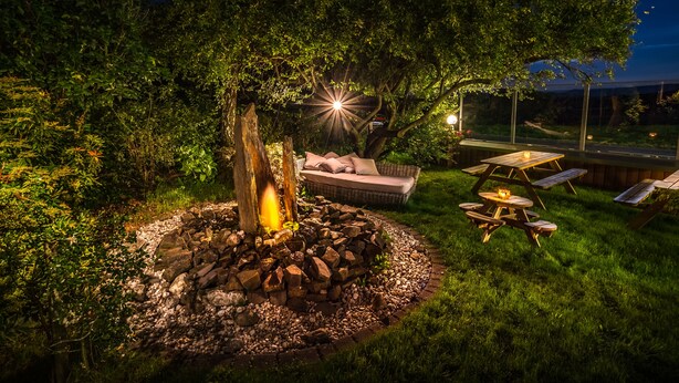 A well-lit outdoor seating area with a stone fire pit, a wicker sofa, and picnic tables. - Hotel Kalenborner Hoehe (Kalenborn)