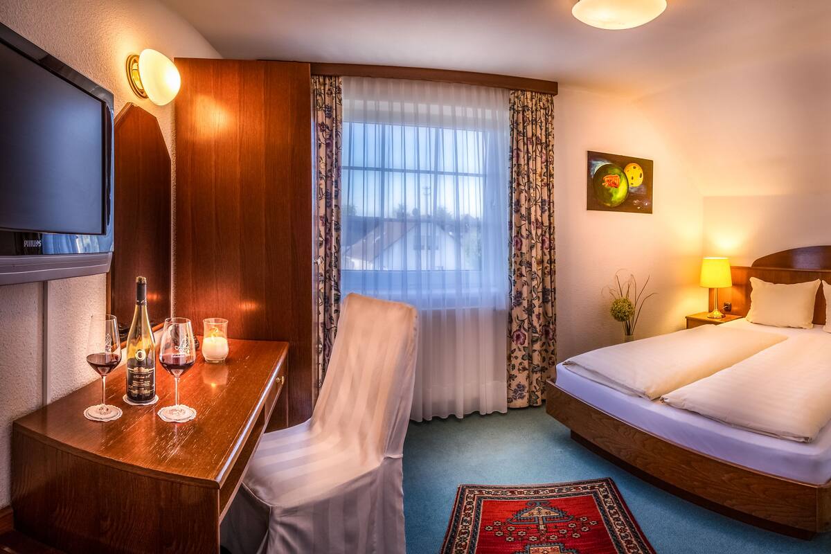 A hotel room with a bed, bedside tables, a TV, a desk with a chair, and a window with curtains.