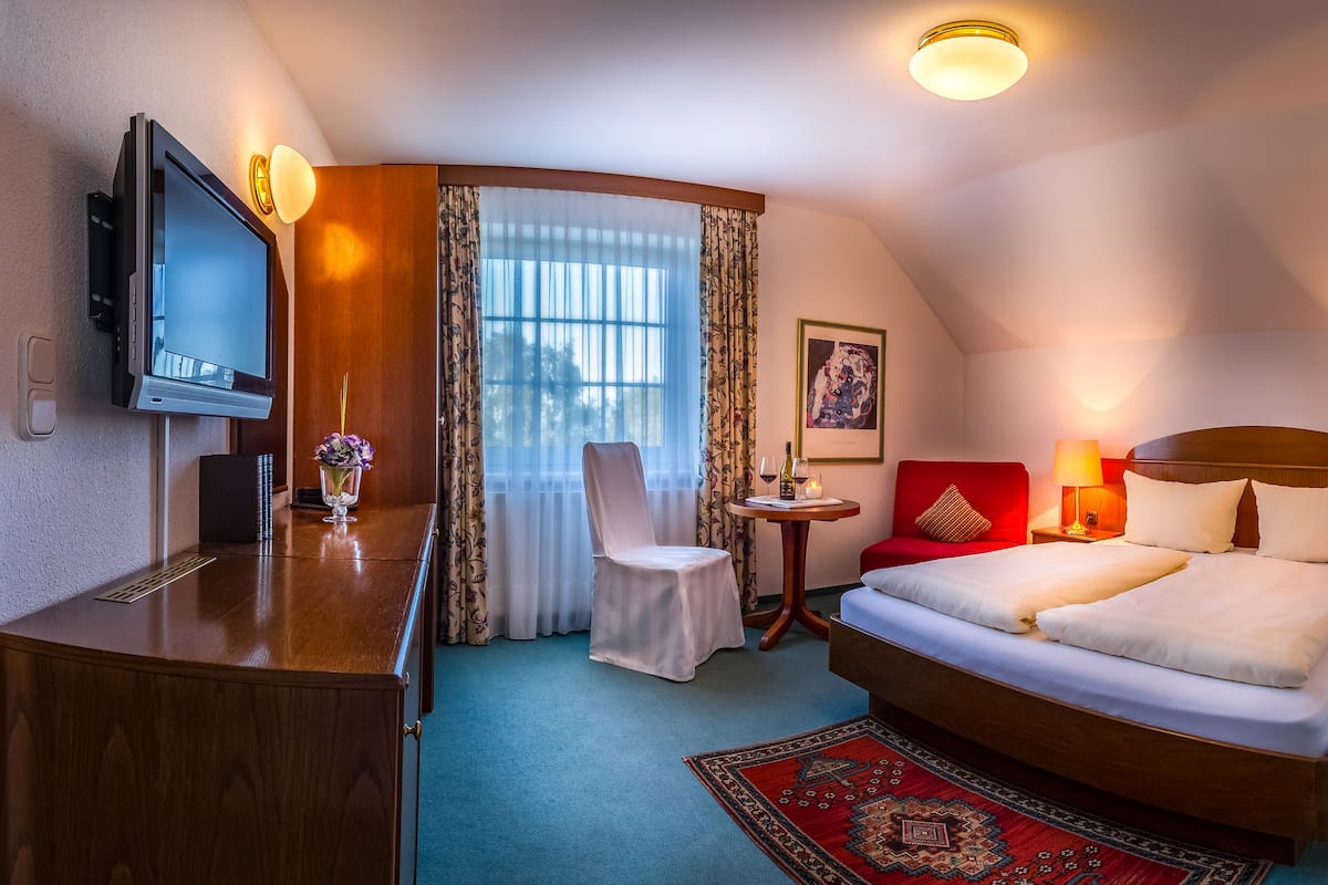 A hotel room with a bed, bedside tables, a desk with a television, a chair, a small table with a candle, and a window with curtains.