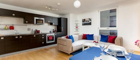 Una cocina moderna con armarios oscuros, un microondas y un hervidor rojo. Hay un sofá beige con cojines azules y rojos, una mesa de comedor con platos y vasos, y una ventana con vistas a la ciudad.