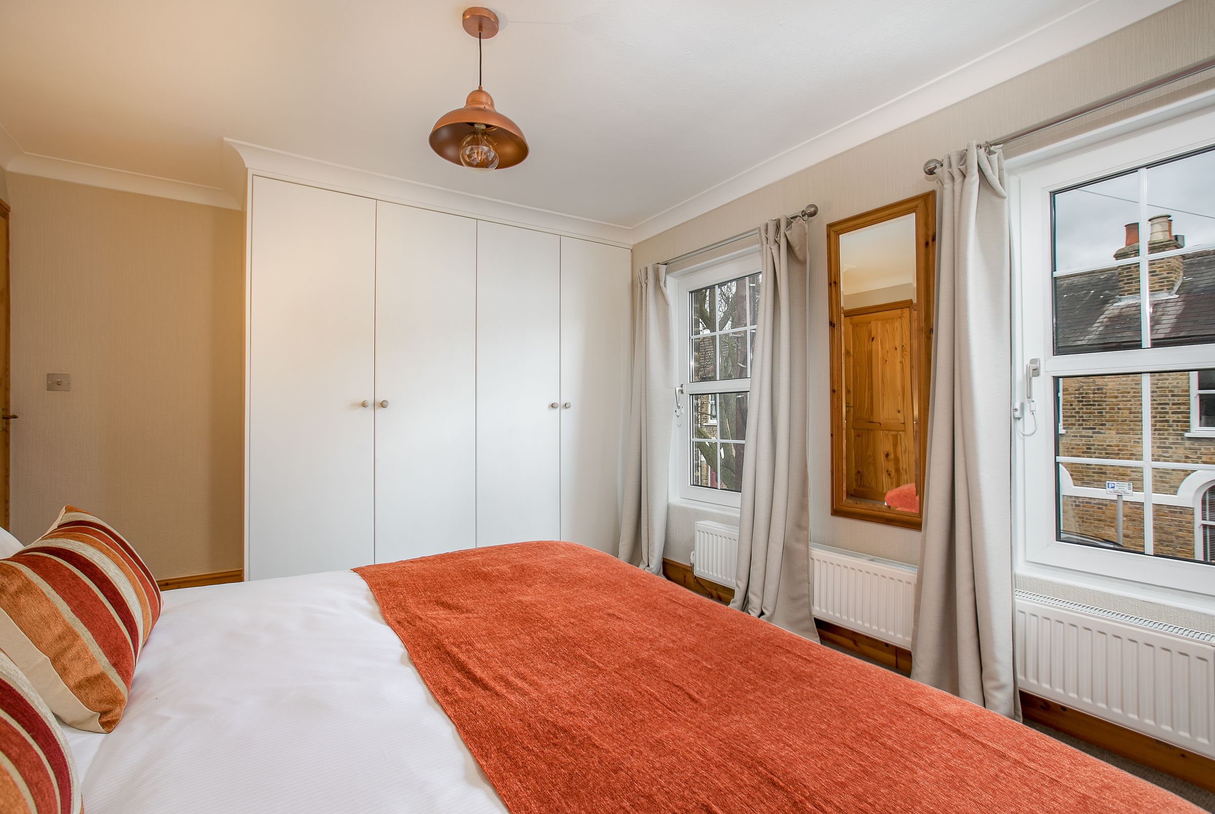 A bedroom with a bed, white wardrobe, large windows with curtains, and a hanging light fixture.