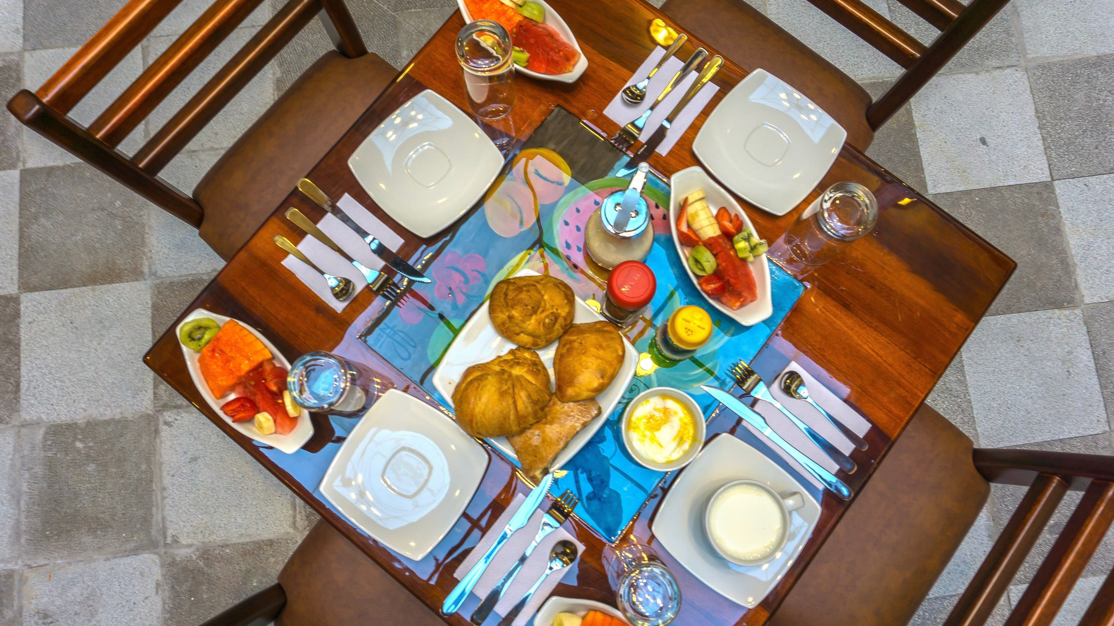 Una mesa de comedor dispuesta con platos, cubiertos y diversos alimentos, incluyendo pasteles y fruta.