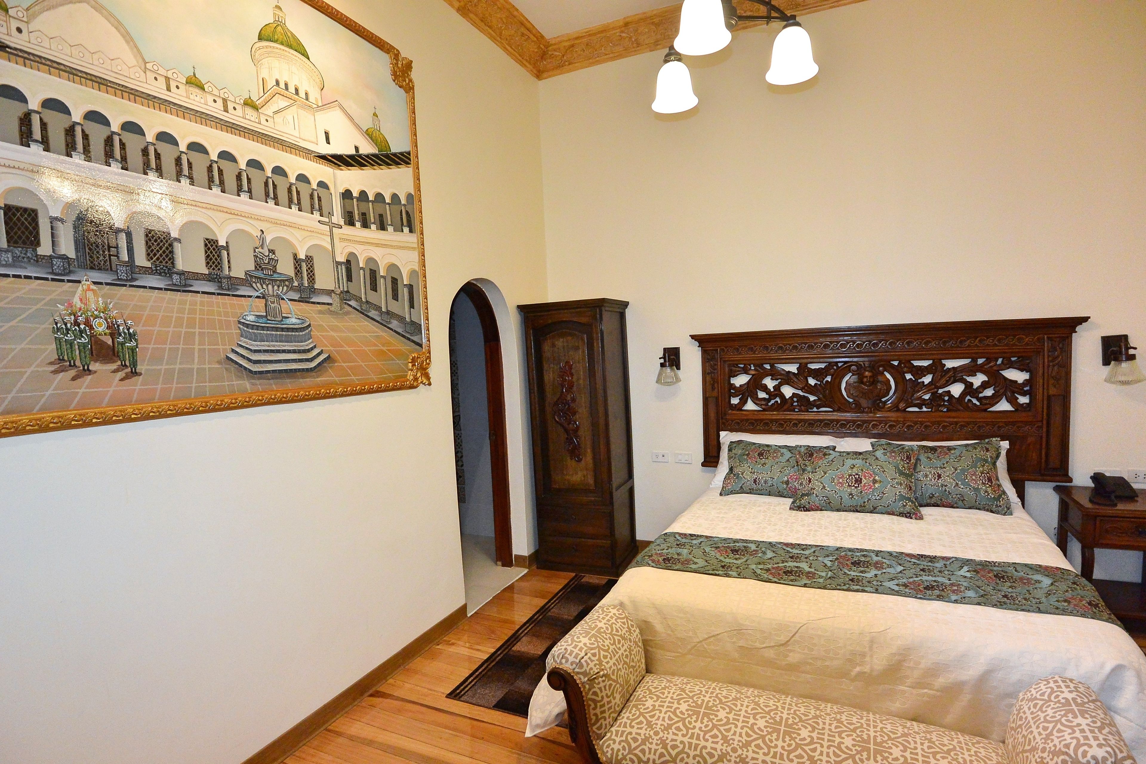 Une chambre à coucher avec un grand lit, une tête de lit en bois, une table de chevet, une commode, un tableau représentant une mosquée et un sol carrelé.