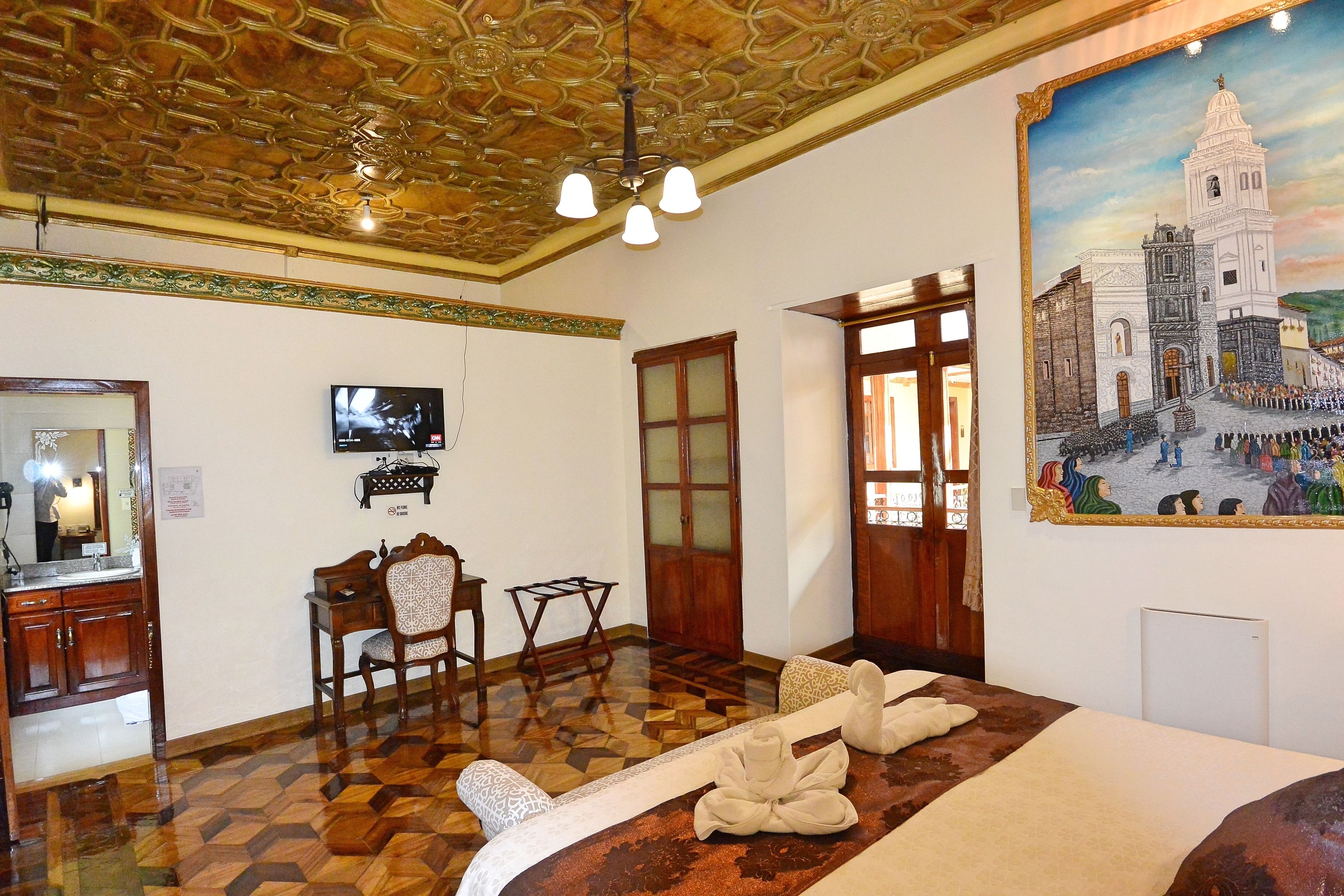 A hotel room with a patterned floor, a bed with towels, a desk with a chair, a television, and a painting of a historical building.