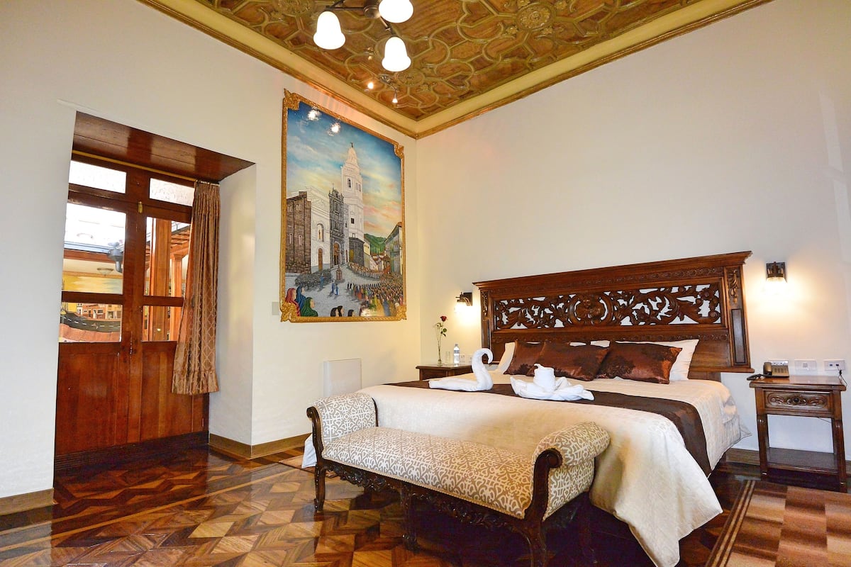 A hotel room with a large bed, a wooden headboard, a painting of a cityscape, a wooden floor, and a nightstand with a lamp.