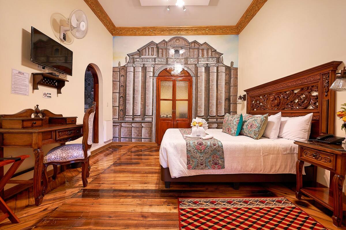 A bedroom with a large bed, a wooden desk, a chair, a television, and a mural of a building on the wall.