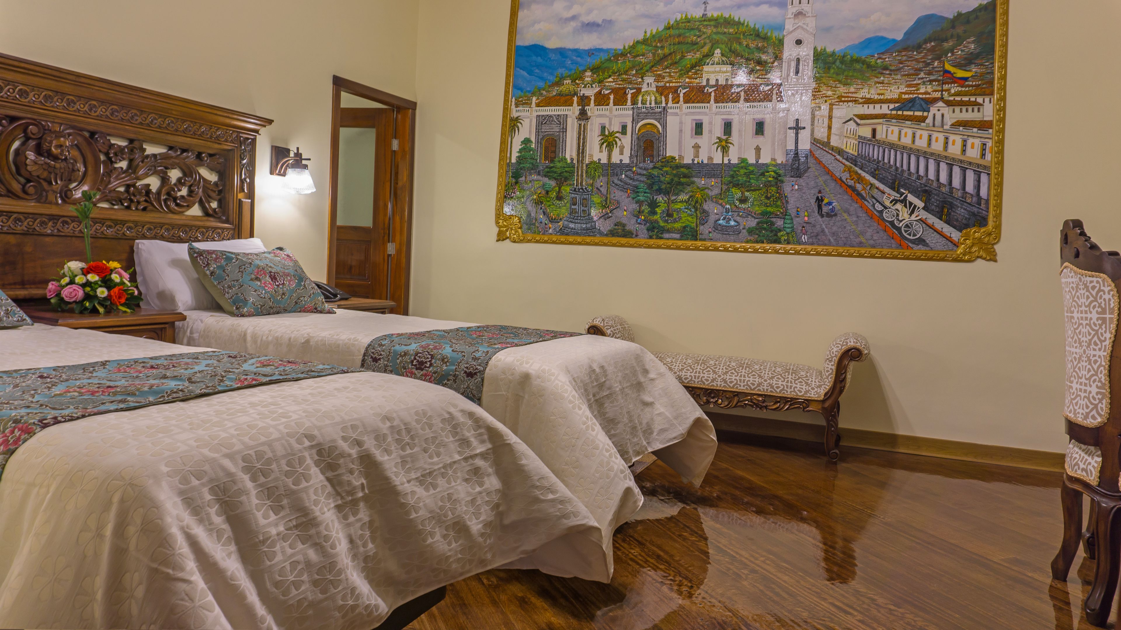 A hotel room with two beds, a wooden headboard with carvings, a bench, and a mural of a cityscape on the wall.