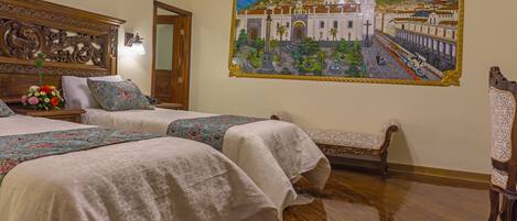 Habitación de hotel con dos camas, cabecero de madera con adornos tallados, un banco y un mural con vista a la ciudad en la pared.