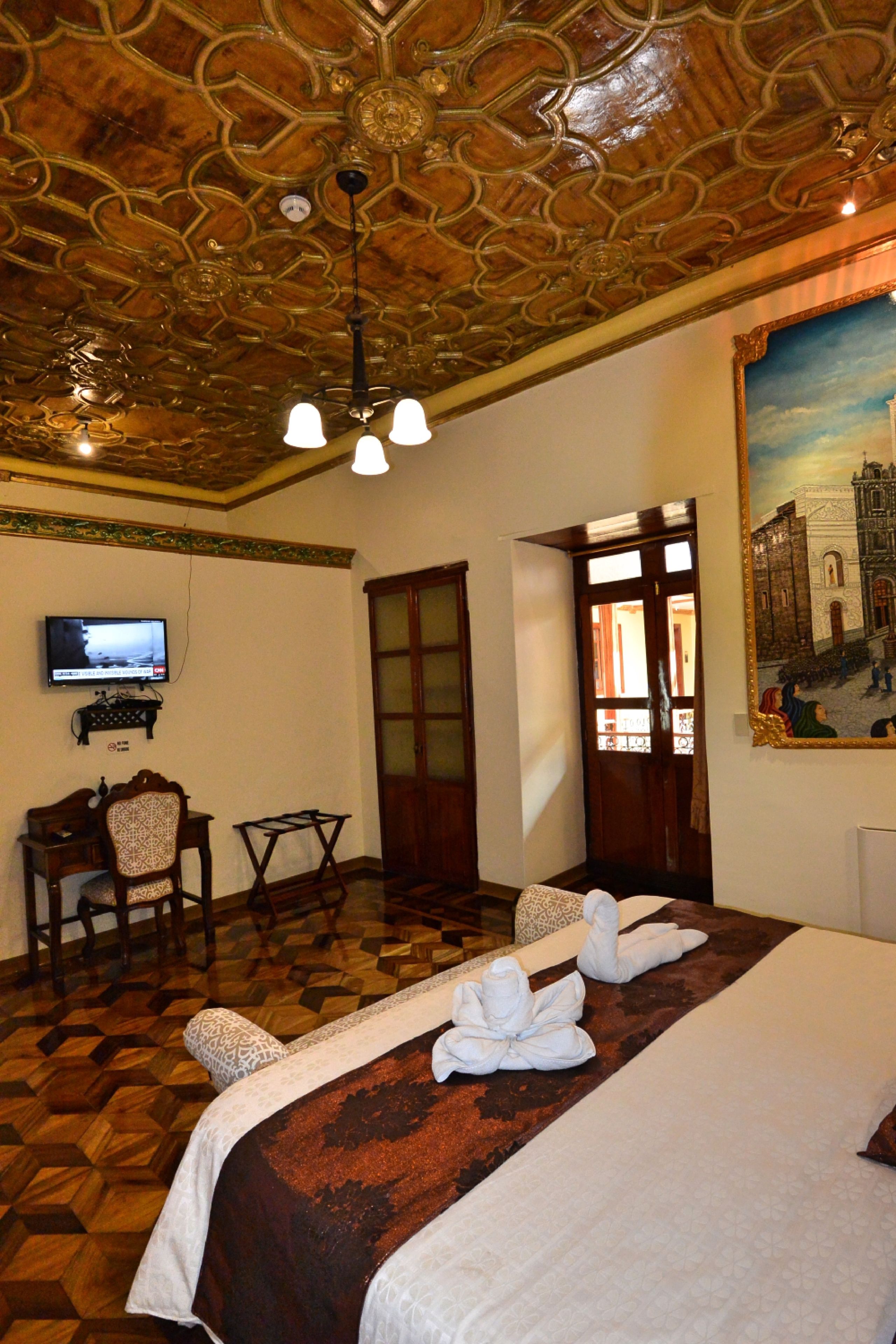A hotel room with a wooden ceiling, a bed with white linens, a desk with a chair, a television, and a painting on the wall.