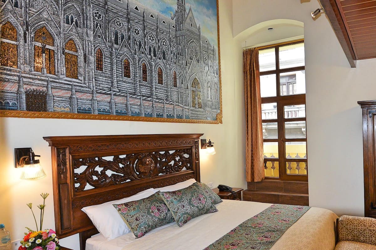 A bedroom with a large bed, a wooden headboard, a nightstand, a lamp, a painting of a cathedral, and a window with curtains.