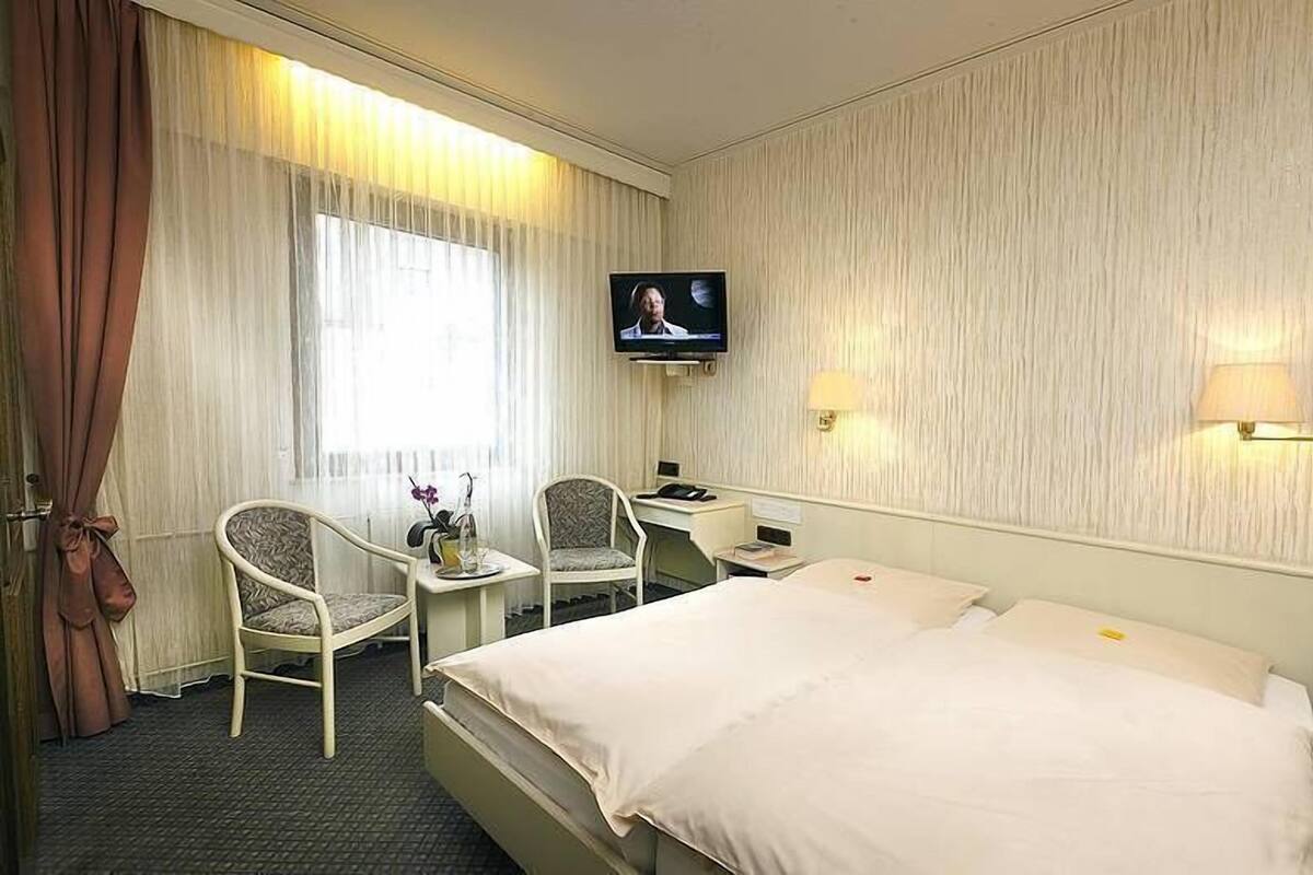 Hotellihuone, jossa on sänky, kaksi tuolia, työpöytä, televisio ja ikkuna, jossa on verhot.