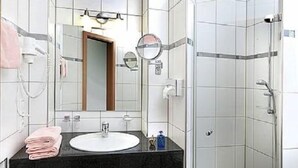 A modern bathroom with a black countertop sink, white tiles, a glass shower enclosure, and a pink rug. - Hotel Pohl zum Rosenberg (Kinheim)