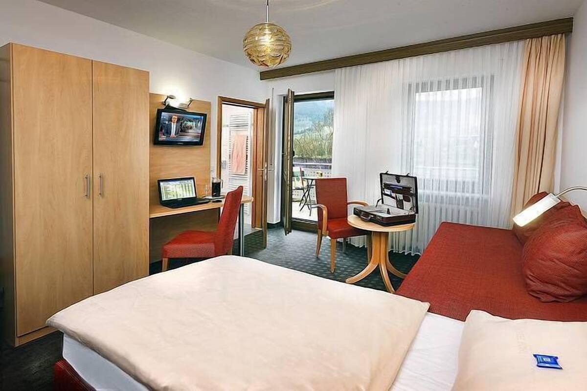 Hotellihuone, jossa on sänky, työpöytä tietokoneella, tuoli, vaatekaappi ja ikkuna, jossa on verhot.