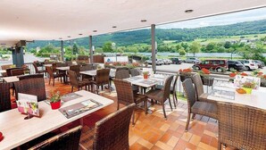 An outdoor seating area with wicker tables and chairs, overlooking a scenic landscape with hills and parked cars. - Hotel Pohl zum Rosenberg (Kinheim)