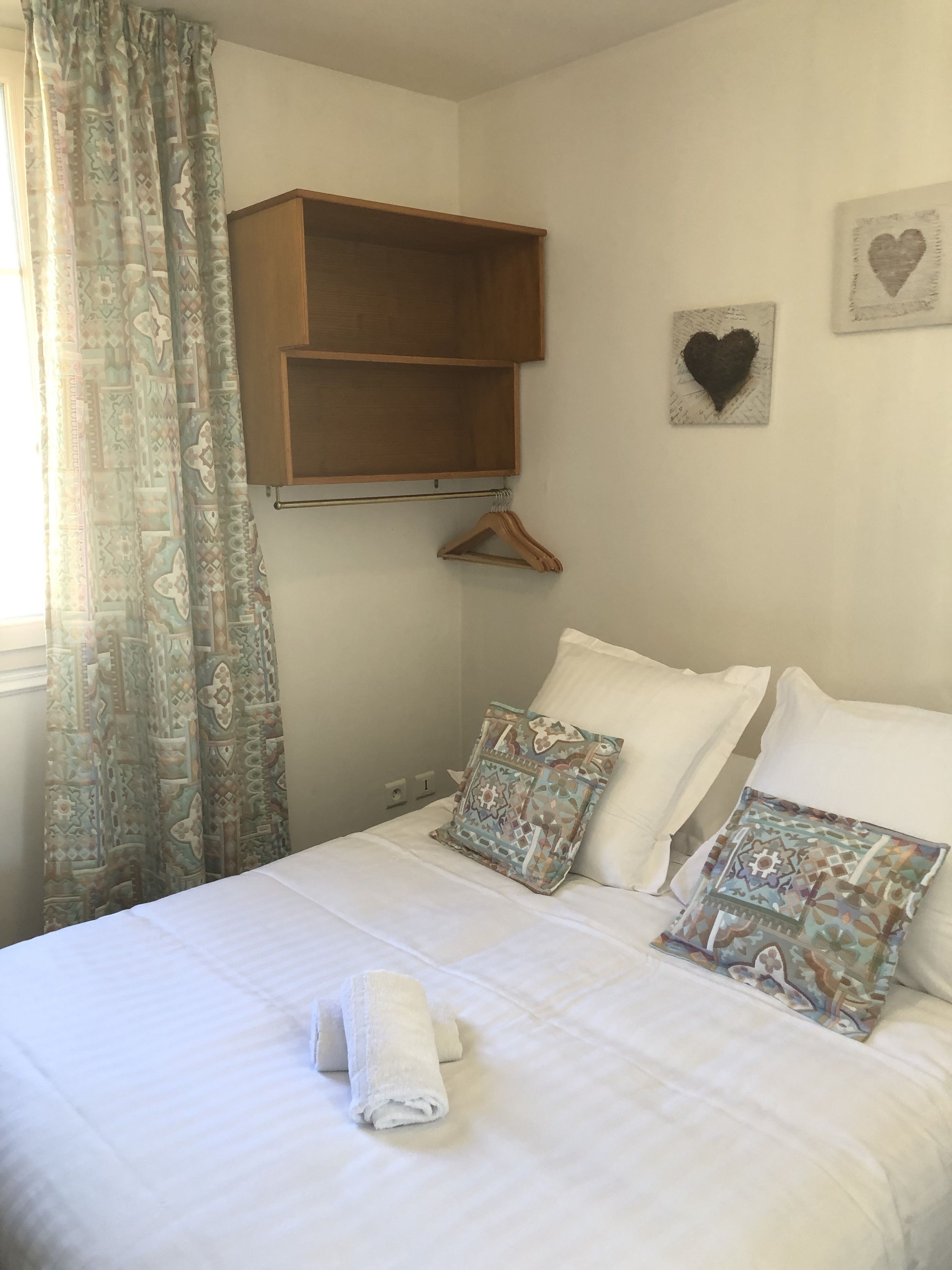 A neatly made bed with two pillows, a hanging wooden shelf, and a wall with a heart-shaped decoration.