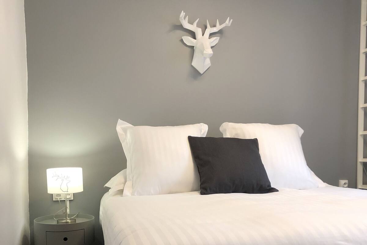 A neatly made bed with white and black pillows, a small bedside table with a lamp, and a deer head decoration on the wall.