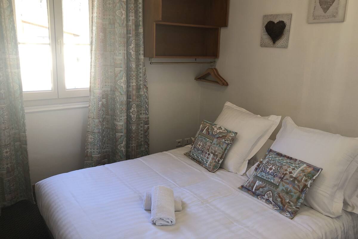 A neatly made bed with patterned pillows, a wooden shelf above, and a window with curtains.