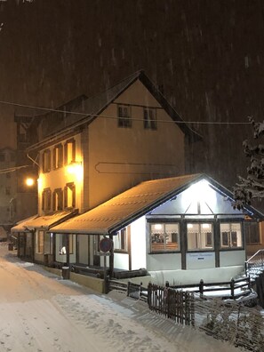 A snow-covered building with a lit entrance, a visible sign, and a wooden fence in front.