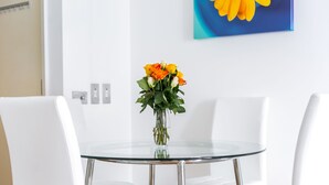 Un tavolo rotondo in vetro con un vaso di fiori, circondato da sedie bianche in una stanza moderna con una parete spoglia e un quadro.