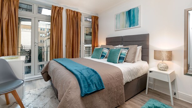 A modern bedroom with a large bed, a bedside table, a chair, and a window with curtains. - Theatreland Walk (London)