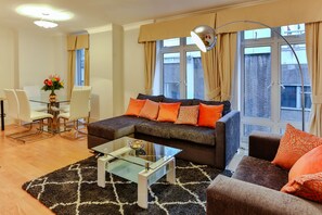 A modern living room with a glass coffee table, a sofa with orange cushions, a dining area with a glass table and chairs, and large windows with curtains.