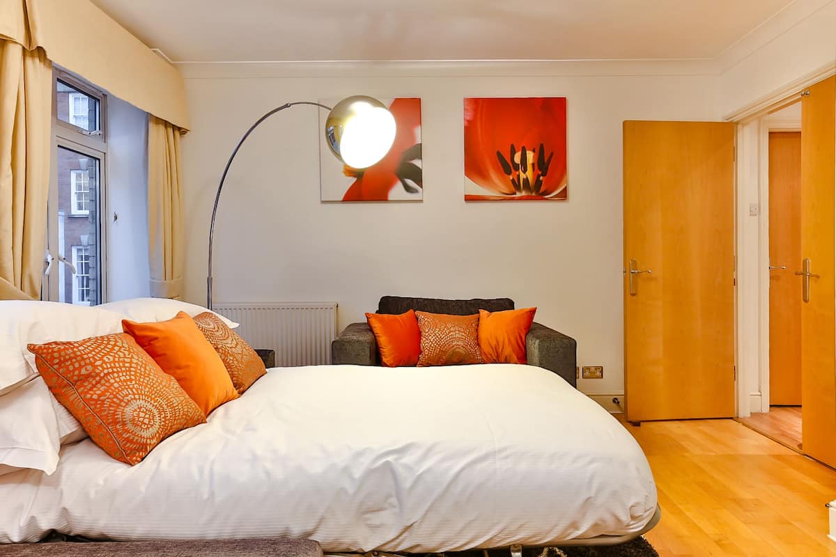 Quarto com uma cama grande, uma escrivaninha e uma cadeira. Há uma janela com cortinas, uma luminária e duas obras de arte na parede.